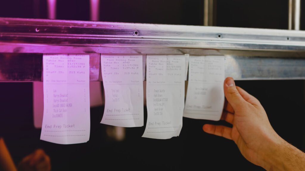 Chef pulling printed order tickets from a kitchen ticket rail inside a restaurant.